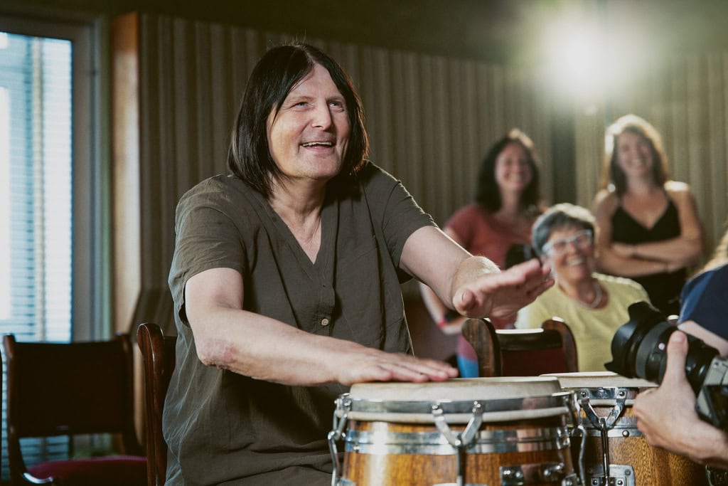 Eine lächelnde Person trommelt entspannt auf Congas, während ein Publikum erfreut zuschaut.