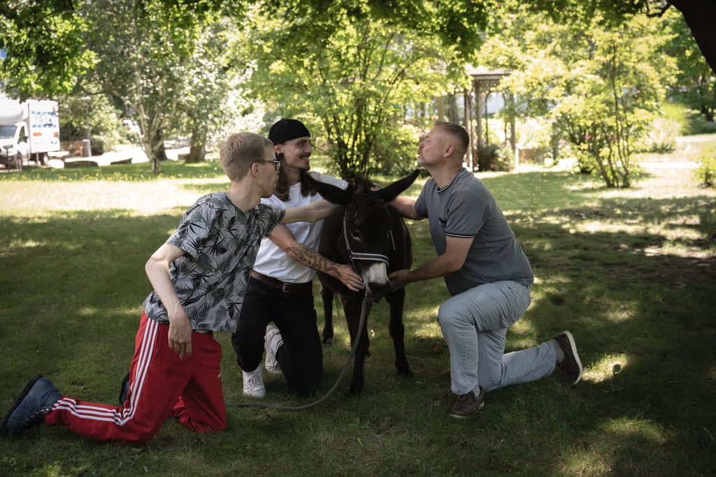 Drei Männer lächeln und knien neben einem Esel im Grünen; einer trägt rote Hosen.
